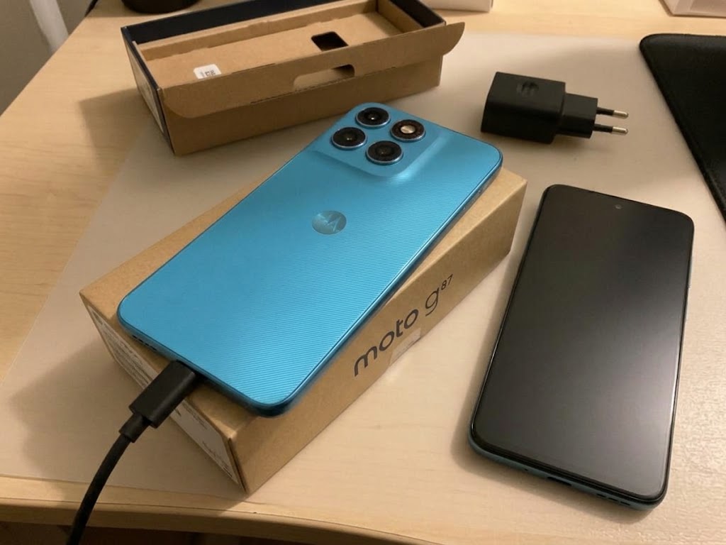 Testowa Motorola Moto G87 w jasnoniebieskiej obudowie z 4 aparatami, podłączona do ładowarki od dołu, leżąca obok pudełka i drugiego telefonu z matowym ekranem